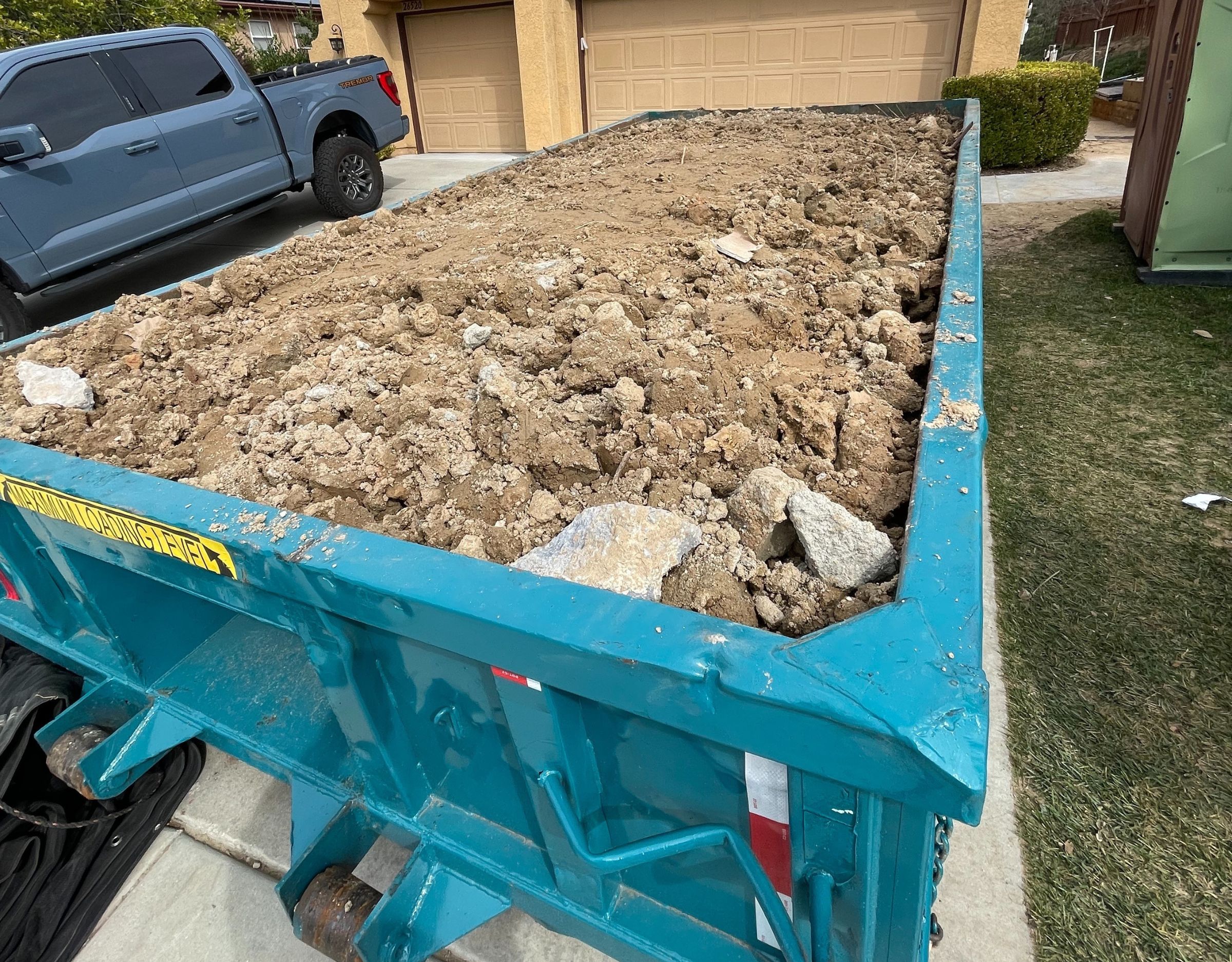 blue 10 yard dumpster in a driveway filed with dirt and other yard waste